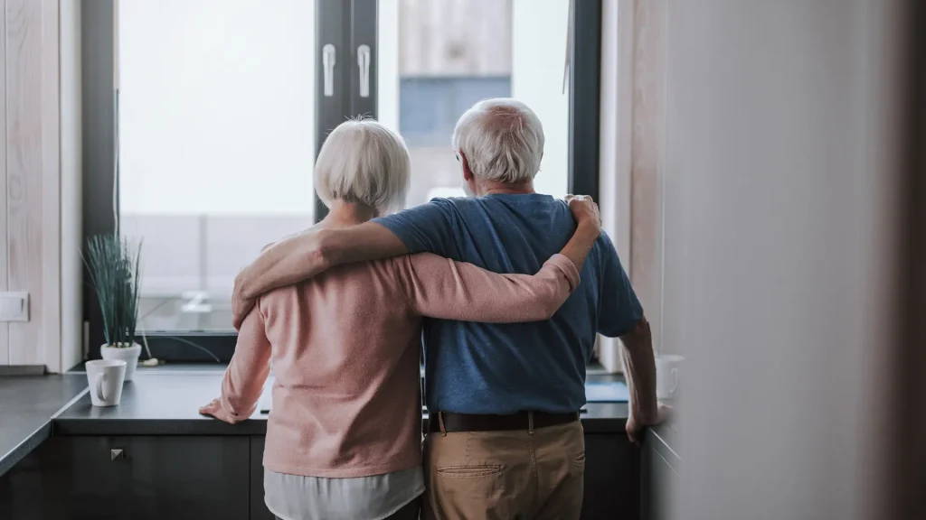 Elderly couple standing by a window with their arms around each other, looking outside.